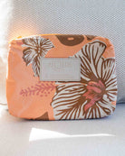 Ein kleines Täschchen aus der „Aloha Collection“ mit einem orange-braunen Blumenmotiv und einem weißen „Aloha“-Etikett wird auf einer strukturierten weißen Oberfläche im strahlenden Sonnenlicht präsentiert. Dieses vielseitige, wasserabweisende Täschchen eignet sich auch hervorragend als stilvolle Bikini-Tasche, um Ihre wichtigsten Utensilien trocken zu halten.