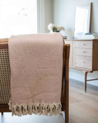 Ein rosa Baumwoll-Plaid „Soft Horizon“ von Main Design mit Fischgrätenmuster und Fransen an den Rändern ist über die Rückenlehne eines Holzstuhls drapiert; im Hintergrund stehen eine Kommode aus hellem Holz und eine Vase mit Blumen.