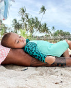 Ein Baby, das einen Kai Baby Badeanzug von Main Design mit Leopardenmuster und UV-Schutz trägt, liegt auf tätowierten Erwachsenenbeinen an einem Sandstrand mit Palmen und einem Pier im Hintergrund unter einem teilweise bewölkten Himmel.