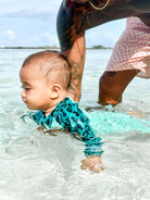 Ein Baby, das den Main Design Kai Baby Badeanzug in blauem Leopardenmuster mit UV-Schutz UPF50+ trägt, wird im flachen Wasser von einem Erwachsenen in rosa gemusterten Shorts unterstützt, dessen tätowierter Arm dem Baby beim Schwimmenlernen hilft.