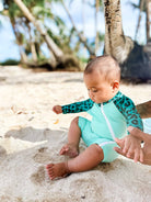 Ein Baby trägt den nachhaltigen Kai Baby Badeanzug von Main Design in Teal und Leo-Print, sitzt am sandigen Strand unter Palmen, spielt mit dem Sand und hält die Hand eines Erwachsenen.