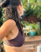 Eine Frau mit nassem, dunklem Haar in einem Dutt steht im Freien und trägt das pflaumenfarbene Sassy Bikini-Top von Main Design. Sie blickt seitlich in die Ferne, während im Hintergrund verschwommene Grünflächen und Terrassenmöbel auftauchen.