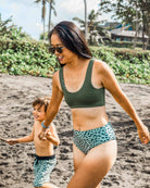 Eine Frau mit Sonnenbrille und einer grünen, leopardenbedruckten Main Design Alice Bikini Hose hält die Hand eines lächelnden Jungen in einer passenden Badehose, während sie an einem palmengesäumten Sandstrand spazieren gehen.