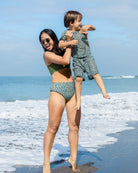 Eine lächelnde Frau mit Sonnenbrille hält ein lachendes Kind am Meer. Beide tragen passende Main Design Alice Bikini-Hosen mit grünen und schwarzen Punkten und stehen auf nassem Sand mit Wellen und blauem Himmel im Hintergrund.