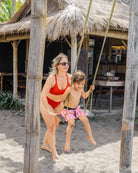 Eine Frau in einer roten Alice Bikini Hose von Main Design schiebt ein Kind auf einer Schaukel an einem sandigen Platz im Freien, umgeben von Bambusstrukturen und einem Strohdachgebäude im Hintergrund.
