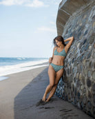 Eine Frau, die den aus recycelten Materialien hergestellten Main Design Serene Surf Bikini Hose trägt, lehnt an einer Steinmauer an einem Sandstrand mit Wellen und dem Meer unter einem teilweise bewölkten Himmel.
