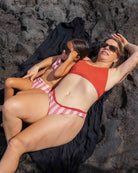 Eine Frau und ein junges Mädchen entspannen sich auf einem schwarzen Handtuch auf dunklem Sand. Beide tragen eine Sonnenbrille und passende Luxe Bikini Hose Badeanzüge von Main Design.