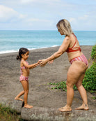 Eine Frau und ein junges Mädchen in passenden Luxe Bikini-Hosen von Main Design halten sich an einem Sandstrand an den Händen, wobei die Frau im Sand liegt und das Mädchen auf einem Baumstamm balanciert, während im Hintergrund das Meer und Grün zu sehen sind.