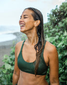 Eine Frau, die das Charm Surf Bikini Oberteil von Main Design trägt, steht im Freien und lächelt unter fallendem Wasser, mit nassen Haaren, üppigen grünen Pflanzen im Hintergrund und einem fernen Strand im Blick.