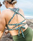 Eine Frau mit hochgestecktem Haar und einer Tätowierung auf dem oberen Rücken sitzt im Sand mit Blick auf das Meer, trägt grüne Leggings und ein gepunktetes Main Design Charm Surf Bikini Oberteil.