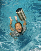 Ein lächelndes Kind in einem gemusterten Badeanzug schwimmt mit einem gestreiften Schwimmer, während der Dry Bag von Meraki Supply Co. by Main Design - ein farbenfrohes, wasserdichtes Outdoor-Accessoire - sicher am Beckenrand liegt.