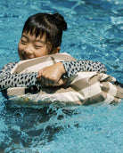 Ein lächelndes Kind in einem gemusterten langärmeligen Badeshirt umarmt einen gestreiften Poolschwimmer und hält seinen Dry Bag von Meraki Supply Co. by Main Design nahe bei sich, während es in einem hellblauen Swimmingpool schwimmt.