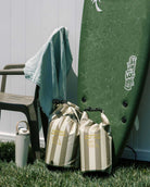 Ein grünes Surfbrett steht auf dem Gras neben einem beigen Stuhl mit einem Handtuch. Zwei Main Design Dry Bag von Meraki Supply Co. und eine wiederverwendbare Wasserflasche warten auf Ihr nächstes Outdoor-Abenteuer, alles vor einem weißen Zaun.