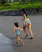 Ein Erwachsener und ein Kind in passenden Lena Mädchen Badeanzügen von Main Design spazieren Hand in Hand an einem Sandstrand entlang in Richtung Wasser, während im Hintergrund Grün und Bäume zu sehen sind.