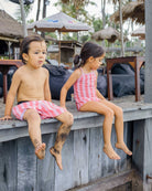 Zwei kleine Kinder in passenden rosa und weißen Badeanzügen, darunter der Lena Mädchen Badeanzug von Main Design, sitzen auf einem Holzbrett am Strand. Der Junge hat sandige Füße, während das Mädchen inmitten von Sonnenschirmen und Palmen zur Seite schaut.
