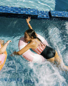 Ein Mädchen in einem rosa-weißen Lena Mädchen Badeanzug von Main Design benutzt beim Spielen im Pool einen passenden Schwimmring aus recycelten Materialien; blaue Fliesen säumen den Rand.