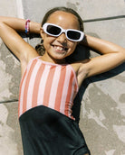 Ein lächelndes Kind in einem Main Design Lena Mädchen Badeanzug und einer weißen Sonnenbrille liegt auf dem nassen Bürgersteig, die Arme hinter dem Kopf, und genießt einen sonnigen Tag.