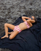 Ein kleines Kind in einem rosa-weiß gestreiften Main Design Nalu Mädchen Bikini mit UPF 50+ Schutz und Sonnenbrille entspannt sich auf einem schwarzen Handtuch auf dunklem Sand, die Hände hinter dem Kopf.