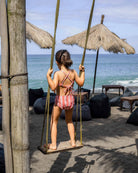 Ein junges Mädchen trägt einen Main Design Nalu Mädchen Bikini und steht auf einer Holzschaukel am Strand. Sie blickt auf den Ozean, während Strohschirme, Sitzsäcke und Tische den Sandstrand unter einem teilweise bewölkten Himmel säumen.