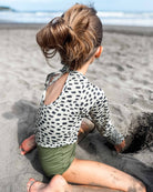 Ein kleines Kind mit hellbraunem Haar in einem unordentlichen Dutt sitzt am Sandstrand, trägt die Kaia Mädchen Badeanzug von Main Design mit UPF 50+ Schutz und gräbt ein Loch im Sand.