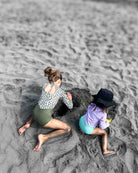 Zwei kleine Kinder graben im Strandsand. Das eine trägt den Main Design Kaia Mädchen Badeanzug mit UPF 50+ Schutz, das andere einen Hut und ein lila Shirt. Beide haben sandige Beine und sind auf ihre Tätigkeit konzentriert.