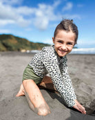 Ein junges Mädchen mit braunen Haaren, das die Main Design Kaia Mädchen Badeanzug und grüne Shorts trägt, kniet lächelnd an einem Sandstrand mit Hügeln und Meer unter einem blauen Himmel. Der Sonnenschutz UPF 50+ schützt sie den ganzen Tag.