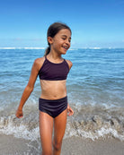 Ein junges Mädchen in einem Nalu Mädchen Bikini von Main Design steht am Rande des Ozeans und lächelt mit geschlossenen Augen, während sanfte Wellen ihre Füße umspülen. Ihr Badeanzug bietet Sonnenschutz mit UPF 50+ unter dem klaren blauen Himmel.