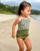 Ein kleines Kind mit nassen Haaren, das den Lena Mädchen Badeanzug von Main Design trägt, lächelt, während es an einem Sandstrand mit Grün und Wasser im Hintergrund steht.