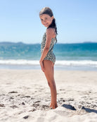 Ein junges Mädchen im Lena Mädchen Badeanzug von Main Design mit verstellbaren Trägern steht barfuß am Sandstrand und lächelt in die Kamera vor einem Hintergrund aus Meer und blauem Himmel.