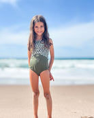 Ein Mädchen lächelt am Strand und trägt den Lena Mädchen Badeanzug von Main Design in Grün-Weiß mit verstellbaren Trägern. Im Hintergrund rauschen Wellen unter einem blauem Himmel mit leichten Wolken.