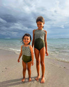 Zwei Mädchen stehen am Strand vor sanften Wellen und bewölktem Himmel, beide tragen den Lena Mädchen Badeanzug von Main Design mit verstellbaren Trägern im grünen, gepunkteten Design.