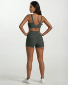 Eine Frau blickt von der Kamera weg und trägt eine dunkelgrüne Main Design Aura Bike Shorts mit mittelhohem Bund und einen passenden Sport-BH. Sie hat lange dunkle Haare, weiße Turnschuhe und steht vor einem schlichten hellen Hintergrund.