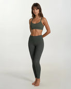 Eine Frau steht barfuß vor einem schlichten, hellen Hintergrund und trägt die Vita Leggings aus recycelten Materialien von Main Design und einen passenden dunkelgrünen Sport-BH. Ihre entspannte Pose unterstreicht die stilvolle nachhaltige Damen Leggings.