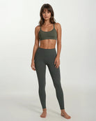 Eine Frau steht barfuß vor einem schlichten Hintergrund. Sie trägt die Vita Leggings von Main Design und einen dazu passenden olivgrünen Sport-BH, die Arme entspannt an den Seiten, der Gesichtsausdruck neutral.