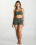 Eine Frau steht barfuß vor einem unifarbenen Hintergrund. Sie trägt das Main Design Essence Active Top und dazu passende hoch taillierte Sportshorts. Sie blickt mit neutralem Gesichtsausdruck und entspannter Körperhaltung nach vorne.