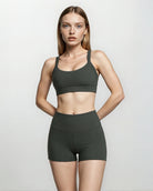 Eine Frau mit glattem blondem Haar steht vor einem schlichten hellen Hintergrund und trägt die Aura Bike Shorts von Main Design in Dunkelgrün mit versteckten Seitentaschen und einen passenden Sport-BH, die Hände hinter dem Rücken, den Blick direkt in die Kamera gerichtet.