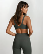 Eine Frau mit langen braunen Haaren trägt die dunkelgrüne Vita Leggings und den passenden Sport-BH von Main Design und steht leicht zur Seite gedreht vor einem schlichten weißen Hintergrund.