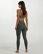 Eine Frau mit langen braunen Haaren steht barfuß mit dem Gesicht zur Wand. Sie trägt die Vita Leggings von Main Design - hochtaillierte, nachhaltige Sportleggings aus recycelten Materialien - und dazu einen passenden dunkelgrünen Sport-BH.