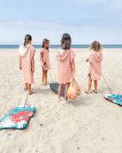 Vier kleine Kinder in passenden rosa Main Design Kinder Badeponcho Flow stehen an einem Sandstrand mit Blick auf den Ozean und halten oder ziehen Strandgegenstände wie ein Surfbrett, eine Tasche und zwei bunte Blumenkissen.