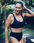 Eine Frau, die die schwarze Alice Bikini Hose von Main Design trägt, lächelt und berührt ihr Haar in einem üppigen grünen Garten, umgeben von tropischen Pflanzen und verschwommenen Outdoor-Möbeln, die eine nachhaltige Sommeratmosphäre schaffen.