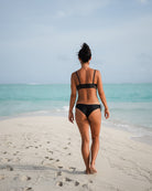 Eine Frau trägt das Sassy Bikini-Top von Main Design, das aus recycelten Materialien hergestellt wurde, während sie an einem Sandstrand steht und auf den türkisfarbenen Ozean unter einem meist klaren Himmel blickt, der eine entspannte tropische Stimmung erzeugt.