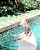 Ein junges blondes Kind sitzt auf einem Surfbrett am Pool, trägt das Main Design Aina Kinder UV Shirt (UPF 50+) und taucht seine Füße ins Wasser, umgeben von üppigen Grünpflanzen und einem Steinweg.