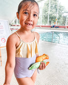 Ein Mädchen trägt den Ava Mädchen Badeanzug von Main Design mit verstellbaren Trägern und steht lächelnd mit nassem Haar am Indoor-Pool, hält bunte Poolspielzeuge. Im Hintergrund sind Schwimmbecken und große Fenster sichtbar.