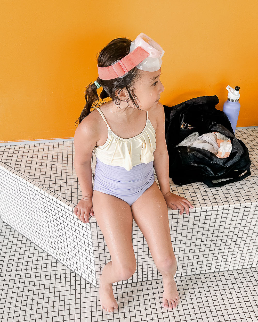 A girl in the Ava Mädchen Badeanzug von Main Design mit verstellbaren Trägern und Schwimmbrille sitzt am Beckenrand. Hinter ihr stehen eine schwarze Tasche und eine lila Trinkflasche vor einer orangefarbenen Wand.