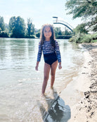 Ein junges Mädchen im Main Design Laila Mädchen Badeanzug mit UPF 50+ geht an einem sonnigen Tag an einem Fluss entlang, mit Bäumen und einer geschwungenen Brücke im Hintergrund.