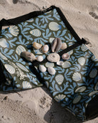 Ein stylischer Dry Bag von Meraki Supply Co. aus dem Hause Main Design mit blau-beigen Muschel- und Korallenmustern liegt im Sand, bestückt mit verschiedenen Muscheln.