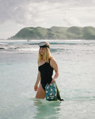 Eine Frau in einem schwarzen Badeanzug steht im flachen Wasser des Ozeans und hält einen Dry Bag von Meraki Supply Co. by Main Design und eine Schnorchelausrüstung. Grüne Hügel und ein wolkenverhangener Himmel bilden eine malerische Kulisse.