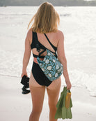 Eine Frau in einem schwarzen Ein-Schulter-Badeanzug steht an einem Sandstrand, hält grüne Schwimmflossen und eine Schnorchelausrüstung in der Hand und hat eine Dry Bag von Meraki Supply Co. by Main Design über die Schulter geworfen, während sie auf das Meer blickt.