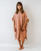 Eine Frau steht barfuß vor einem weißen Hintergrund und trägt den Shoreline Breeze Poncho von Main Design - ein lockeres, strukturiertes Kaftankleid mit kurzen Ärmeln und einer lockeren Passform in Einheitsgröße in Hellbraun oder Pfirsich.