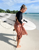 Eine Person mit einem Surfbrett steht an einem Sandstrand und trägt ein handgefertigtes schwarzes Langarmshirt und den rostfarbenen Sarong Bali von Main Design. Das Meer und die Bäume bereichern die Kulisse von Bali unter einem teilweise bewölkten Himmel.